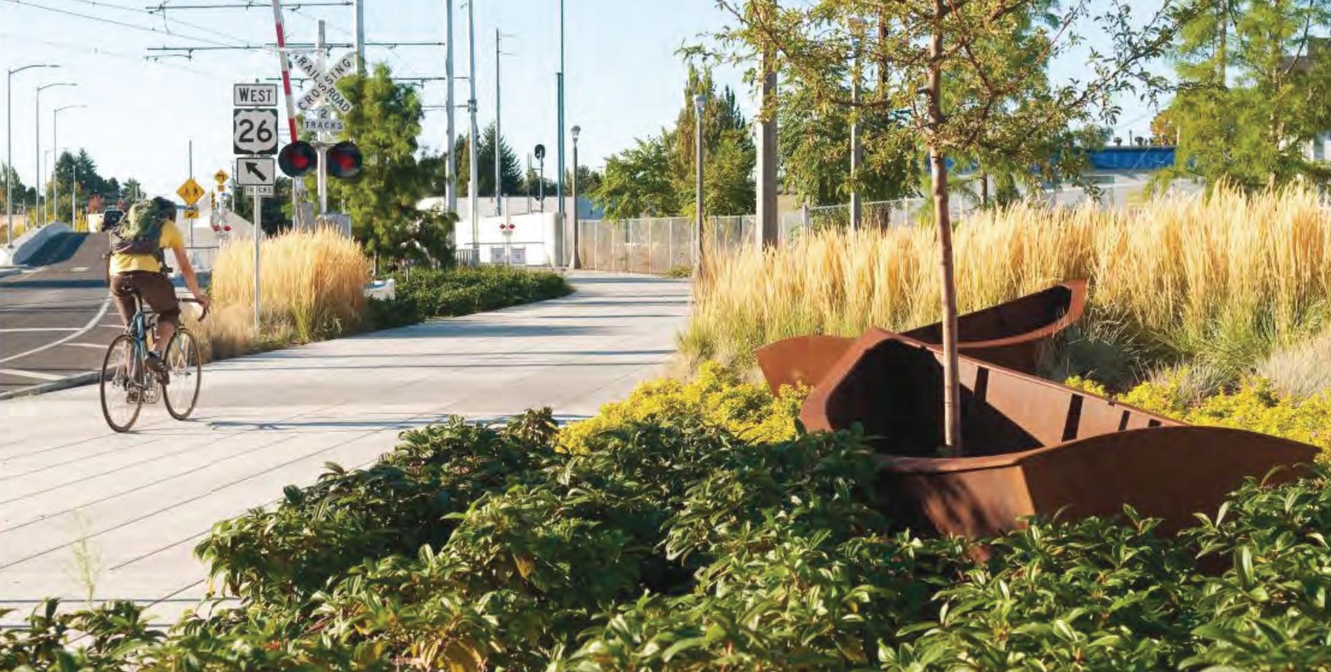 Cyclist riding past native plantings and a boat-like metal art piece along the MAX Orange Line corridor.